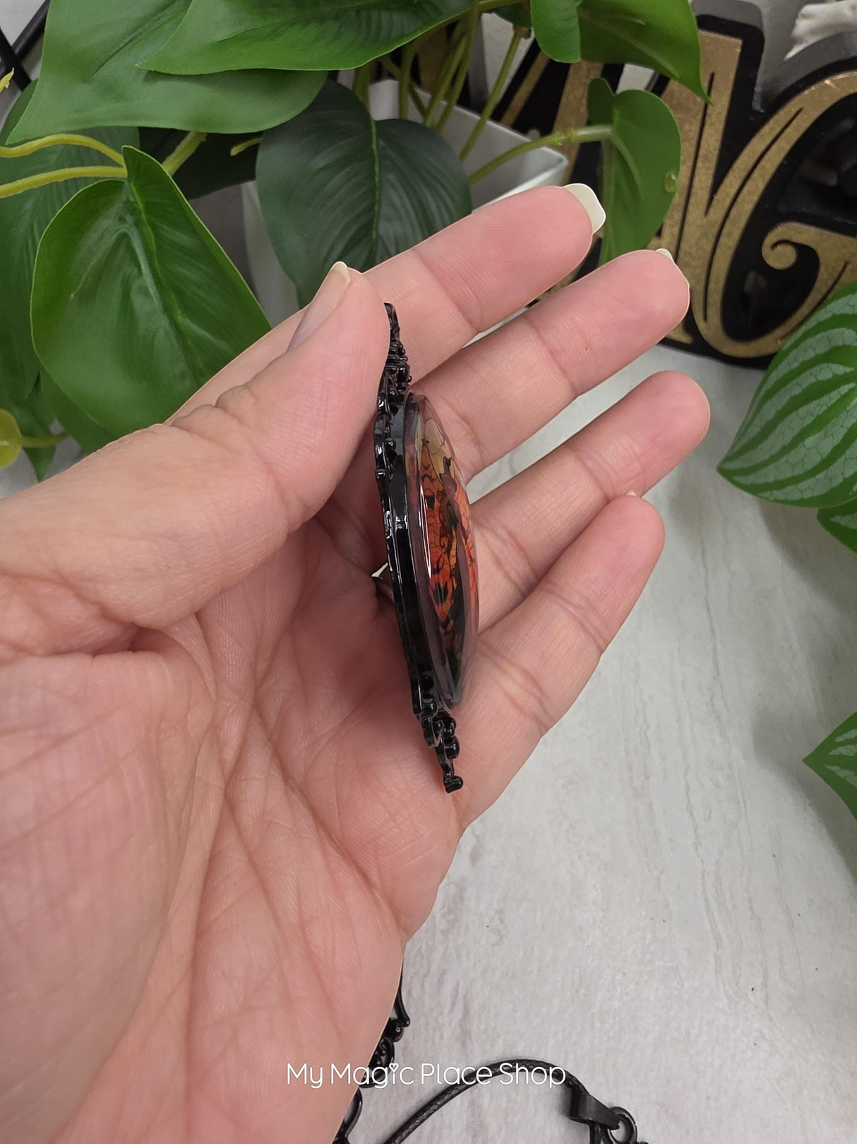 My Magic Place Shop oval witch glass cabochon pendant in an ornate black metal frame, side view held in hand.