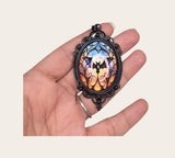 oval gothic bat pendant with a stained glass style cabochon in a black filigree metal frame.
